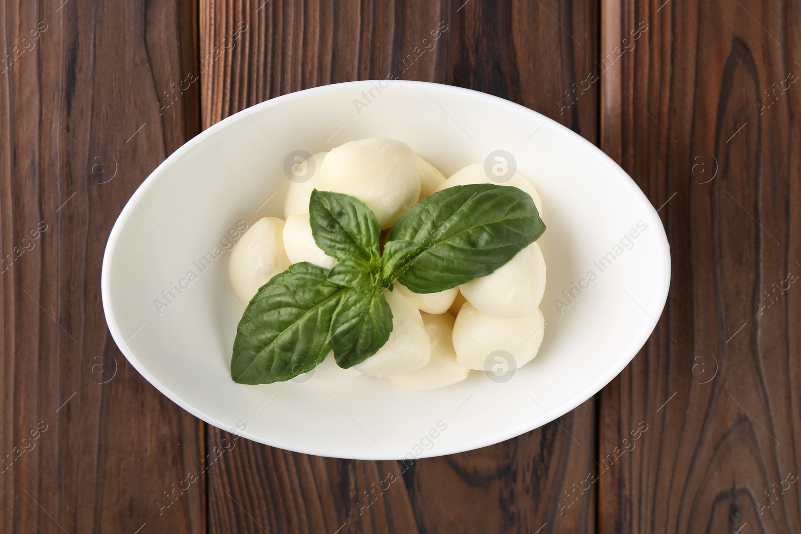 Photo of Tasty mozarella balls and basil leaves in bowl on wooden table, top view