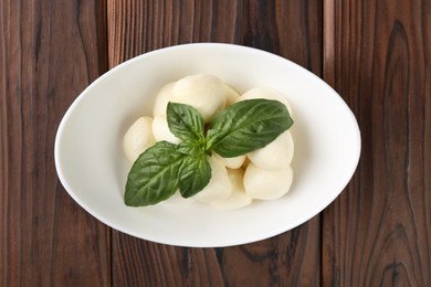 Tasty mozarella balls and basil leaves in bowl on wooden table, top view