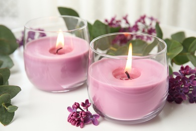 Photo of Burning candles in glass holders and flowers with leaves on white table