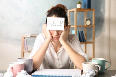 Photo of Young woman with note HELP on forehead at workplace