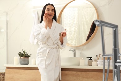 Photo of Beautiful young woman applying cream onto face near mirror in bathroom