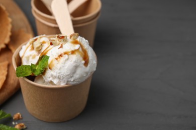 Photo of Tasty ice cream with caramel sauce, mint and nuts in paper cup on grey wooden table, closeup. Space for text