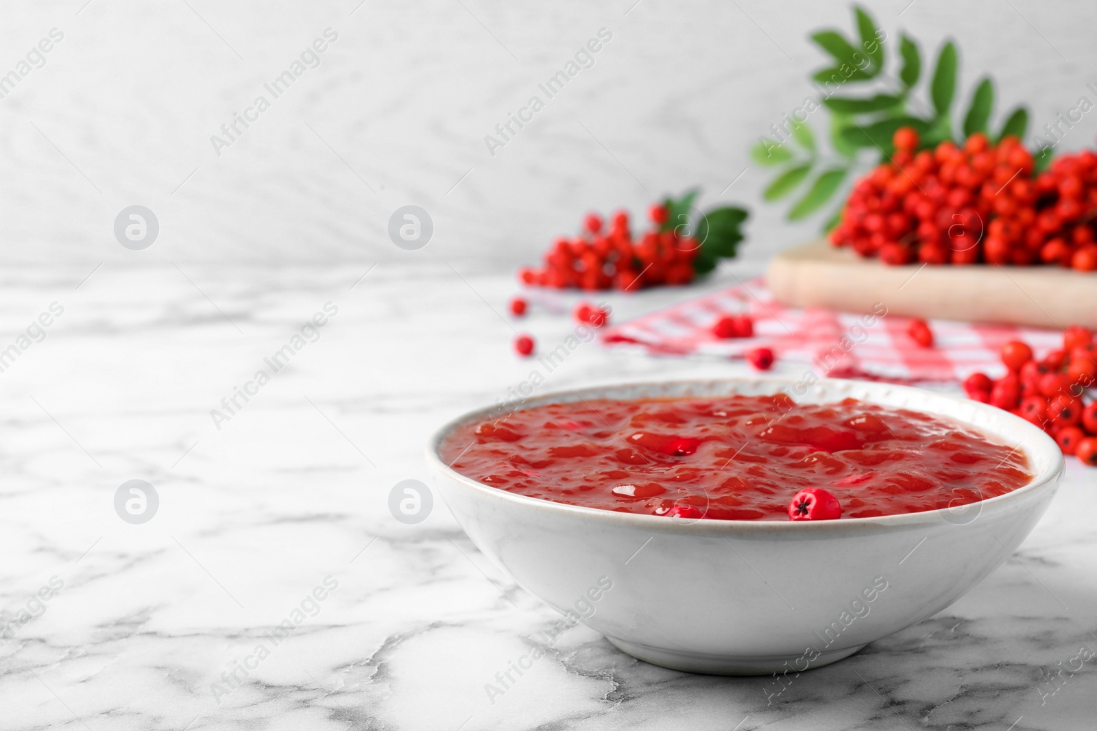 Photo of Delicious rowan jam in bowl and berries on white marble table. Space for text