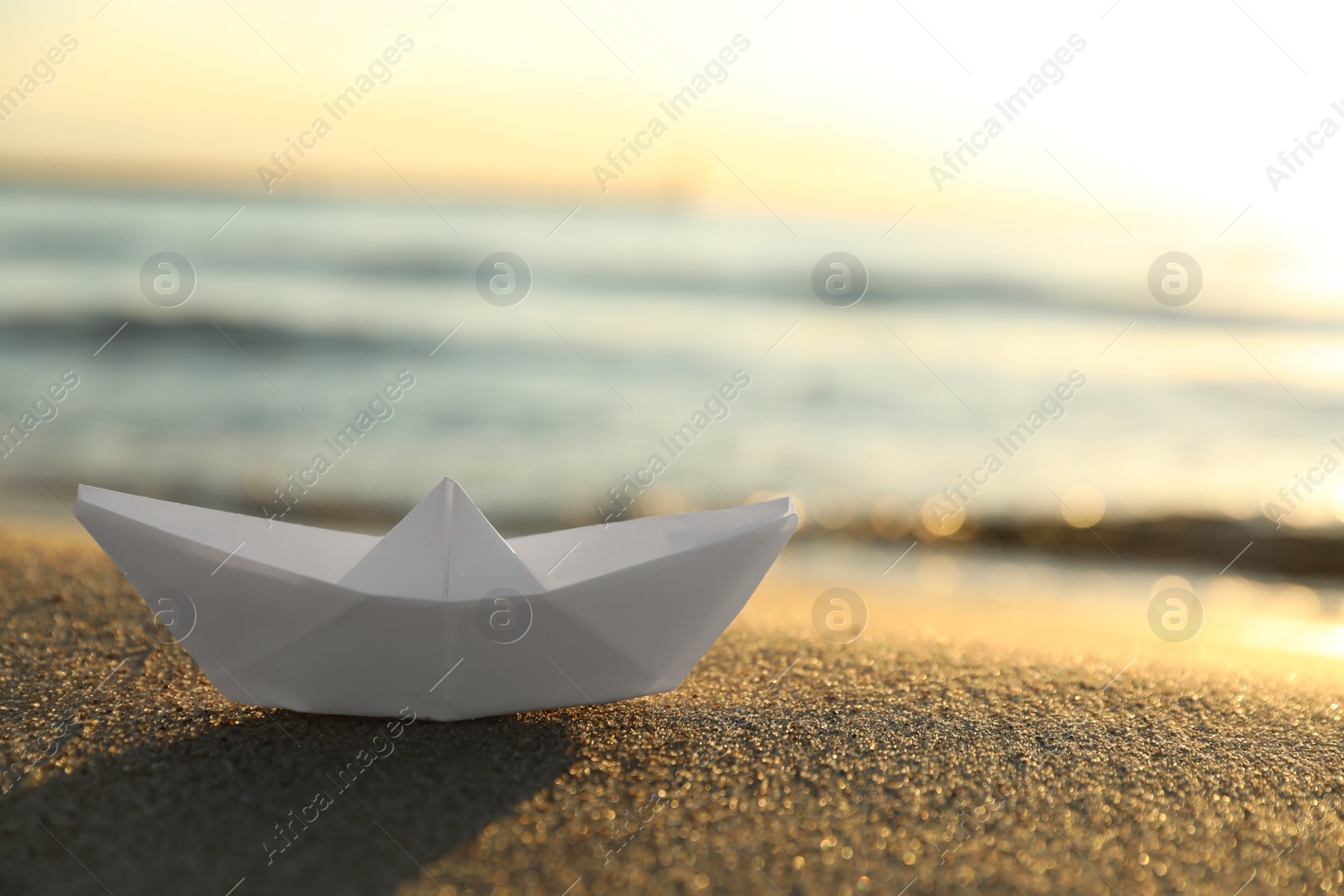 Photo of White paper boat on sand near sea at sunset, closeup. Space for text