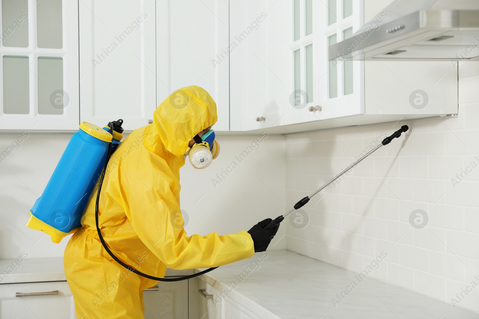 Photo of Pest control worker in protective suit spraying insecticide on furniture indoors