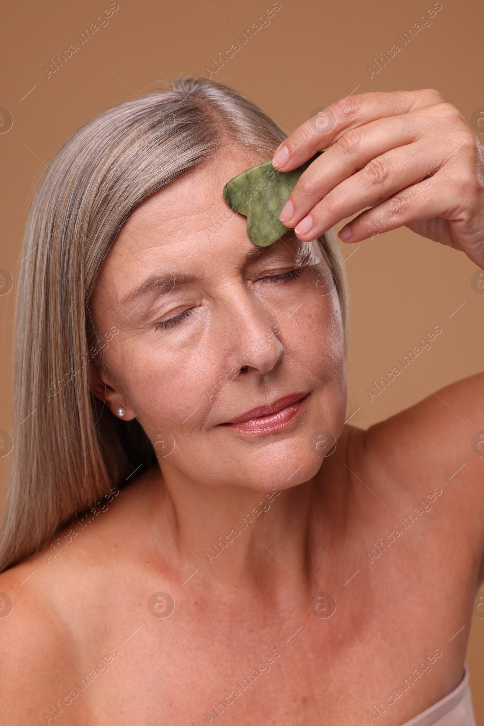 Photo of Woman massaging her face with jade gua sha tool on brown background