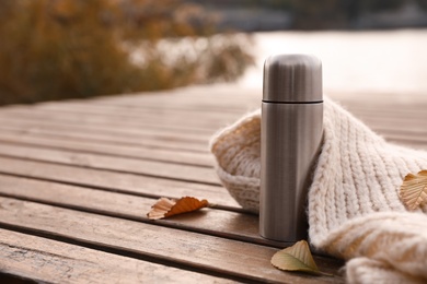 Photo of Metallic thermos, knitted scarf and fallen leaves on wooden pier near river. Space for text