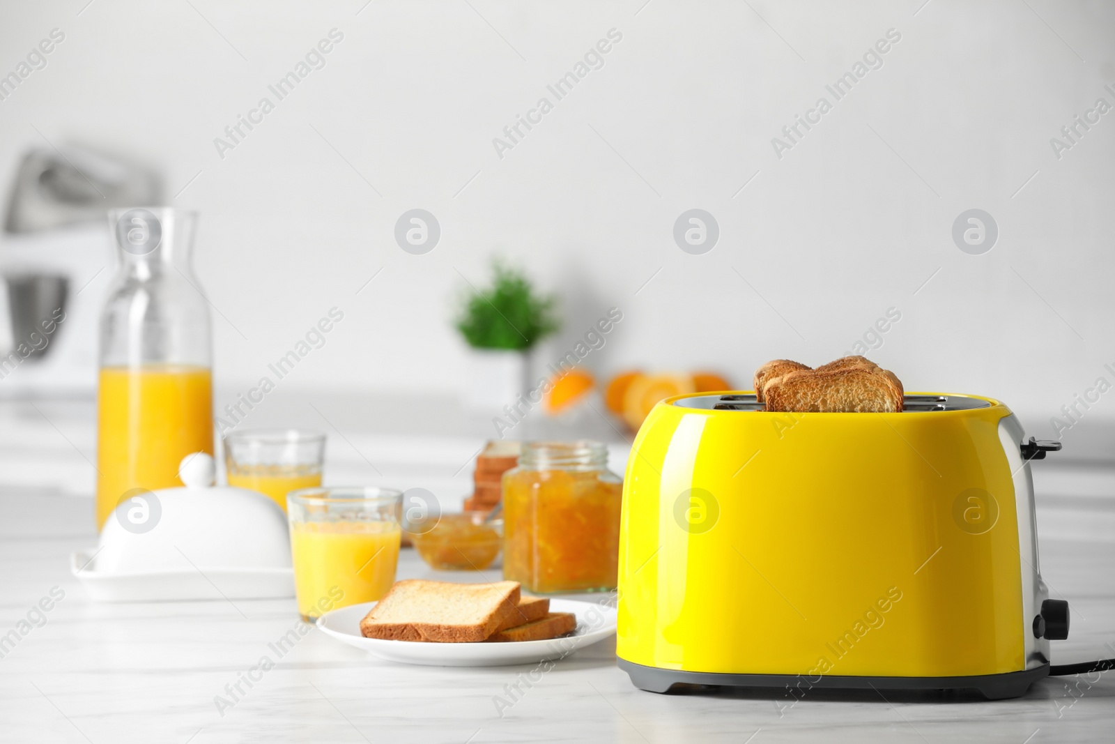 Photo of Yellow toaster with roasted bread slices, jam and glasses of juice on white marble table