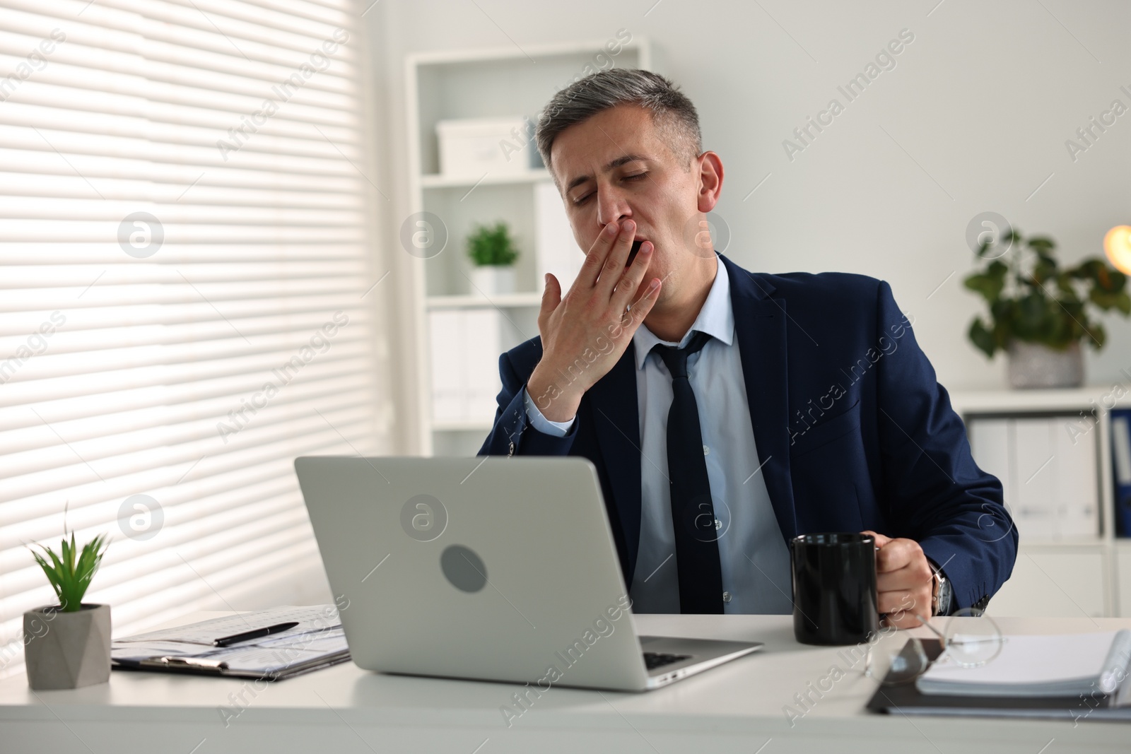 Photo of Sleepy man with cup of drink yawning at table in office