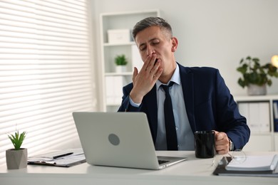 Sleepy man with cup of drink yawning at table in office