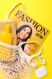 String bag with fashion magazine and beach accessories on yellow background, flat lay