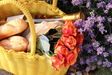 Photo of Yellow wicker bag with beautiful roses, bottle of wine and baguettes near lavender flowers outdoors, above view