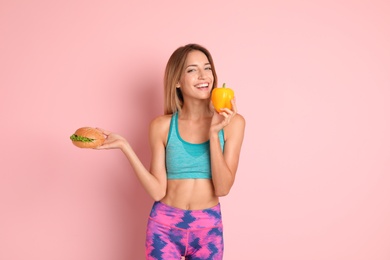 Young woman with burger and bell pepper on color background. Choice between diet and unhealthy food