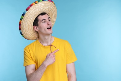 Photo of Young man in Mexican sombrero hat pointing at something on light blue background. Space for text