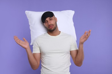 Tired man with pillow and sleep mask on violet background. Insomnia problem