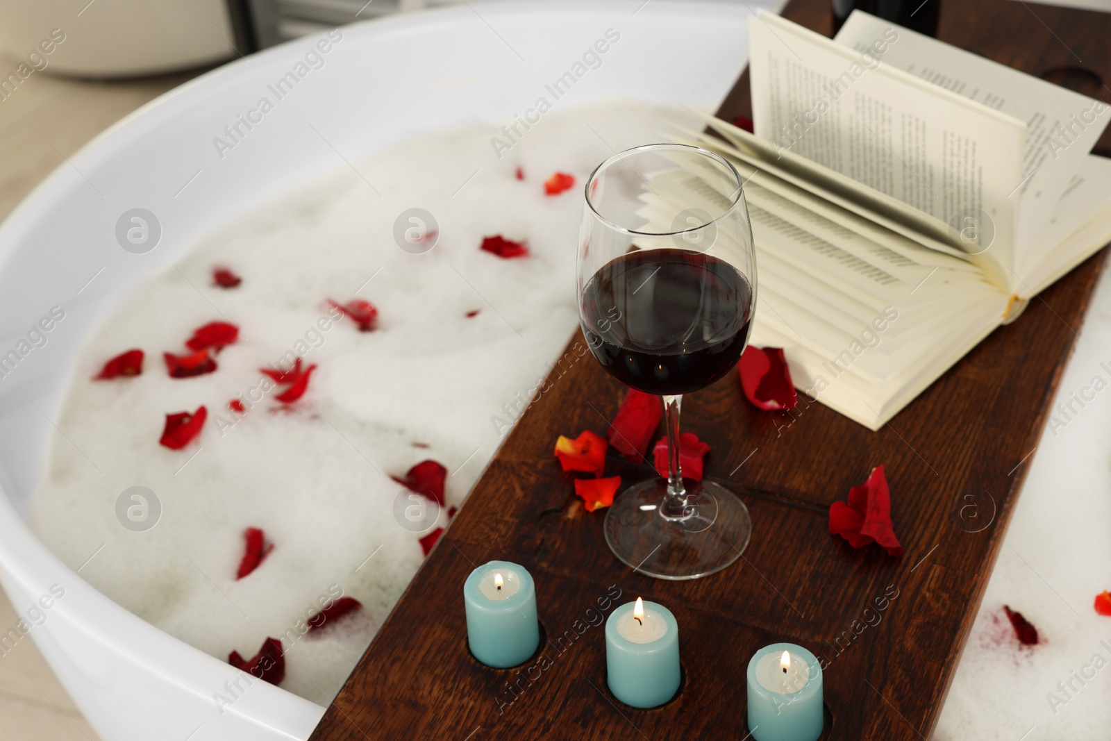 Photo of Wooden board with glass of wine, book, burning candles and rose petals on bath tub