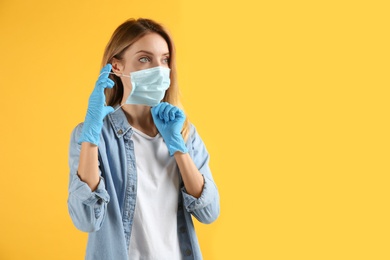 Photo of Young woman in medical gloves putting on protective face mask against yellow background. Space for text