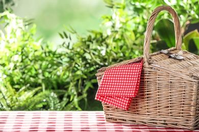 Photo of Closed wicker picnic basket on checkered tablecloth against blurred background, space for text