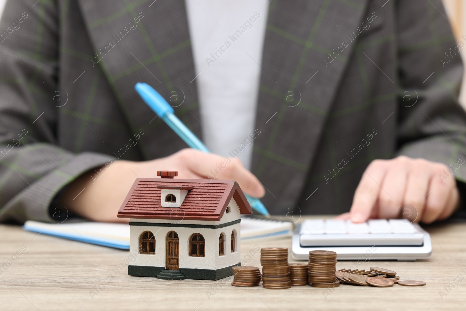 Photo of Woman writing at wooden table, focus on house model and stacked coins