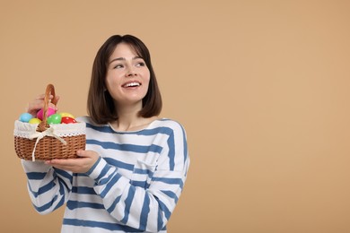 Easter celebration. Happy woman with wicker basket full of painted eggs on beige background, space for text