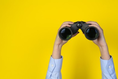 Photo of Woman holding modern binoculars on yellow background, closeup. Space for text