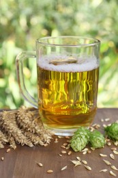 Photo of Mug with beer, fresh hops and ears of wheat on wooden board outdoors