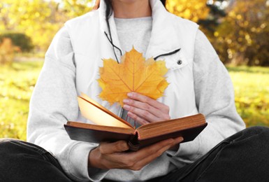 Photo of Woman reading book and holding dry leaves in park on autumn day, closeup