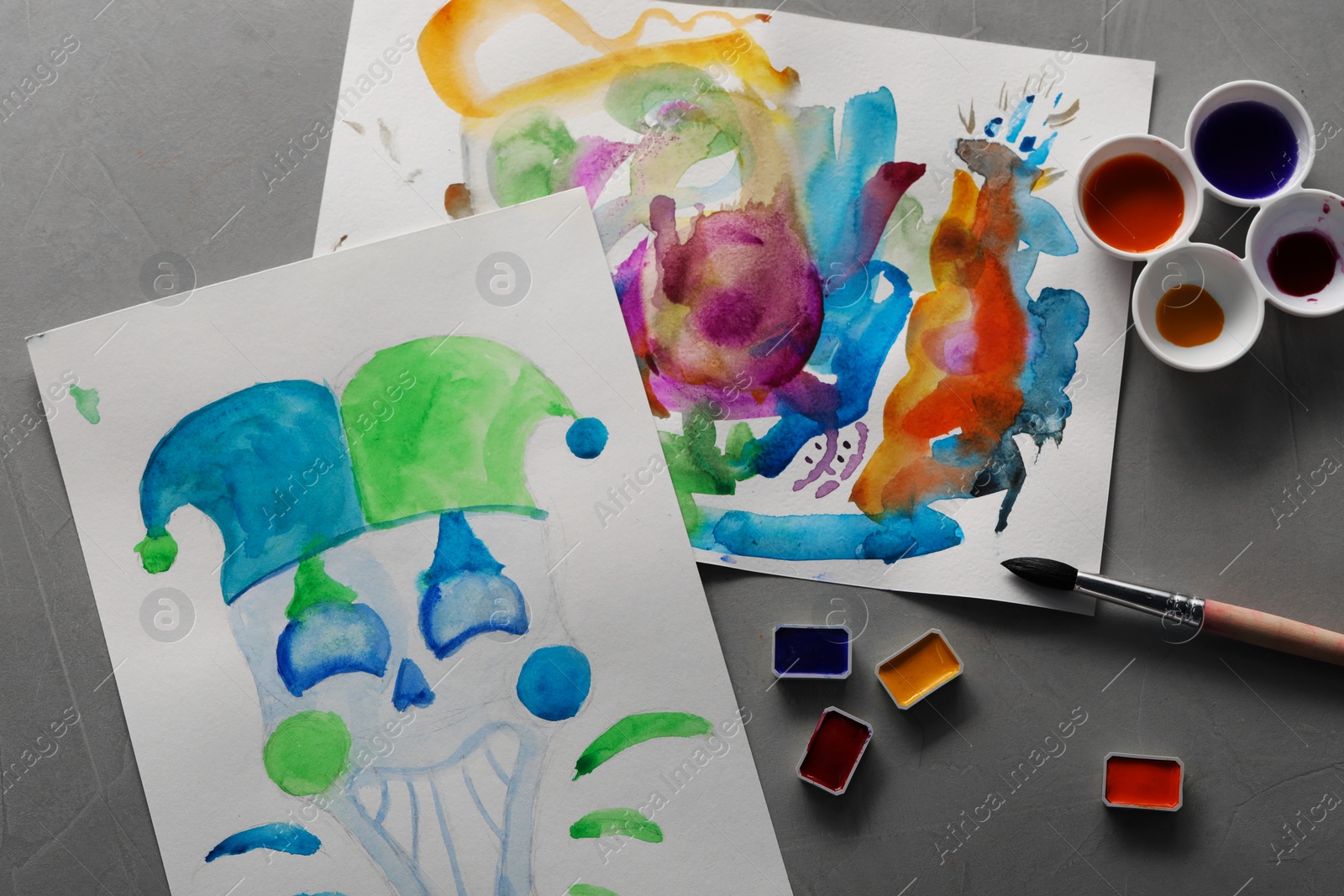 Photo of Sheets of paper with watercolor paintings, brush and paints on grey table, flat lay
