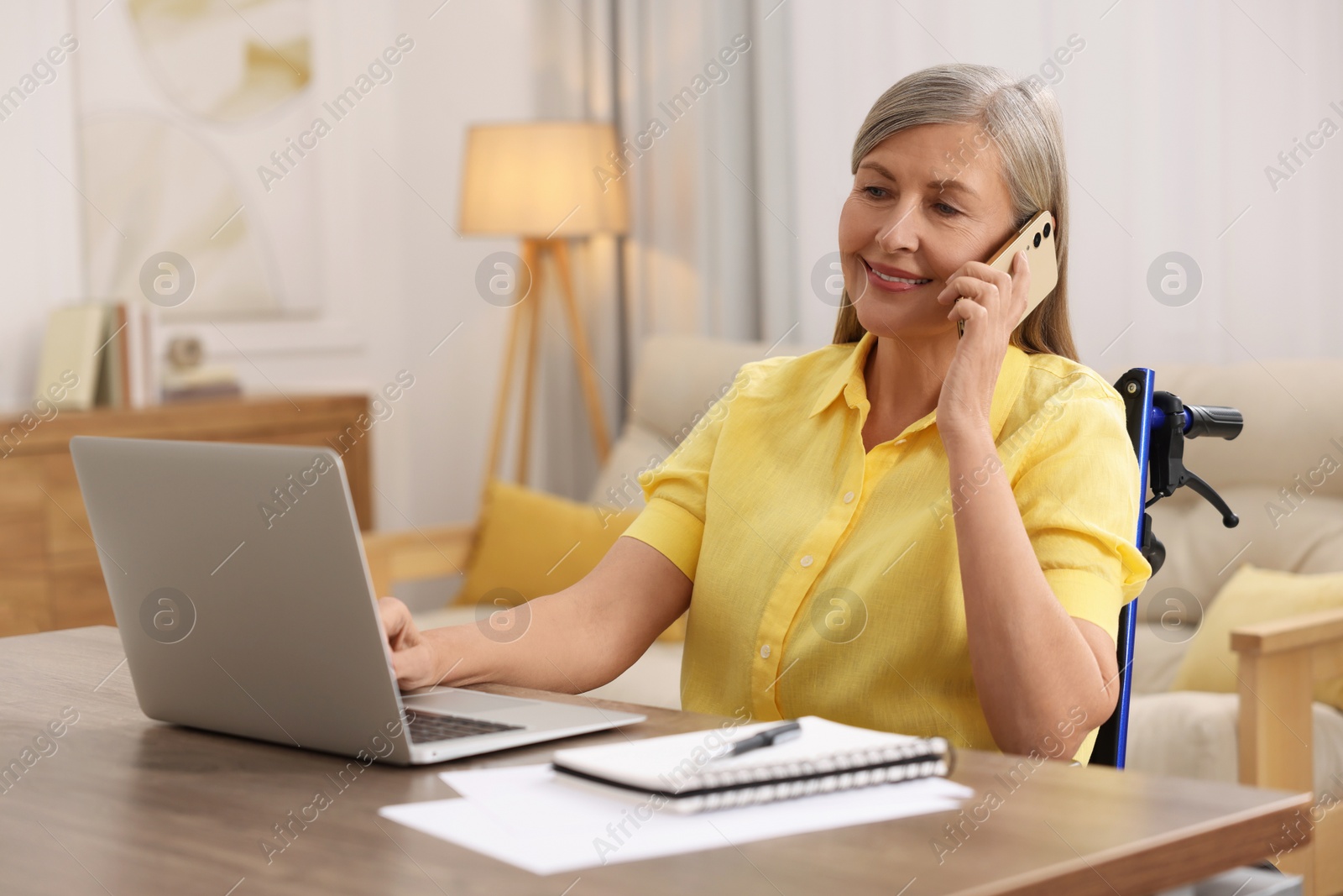 Photo of Woman in wheelchair talking on smartphone while using laptop at table in home office