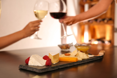 Photo of Different delicious cheeses on table and blurred couple toasting with wine on background