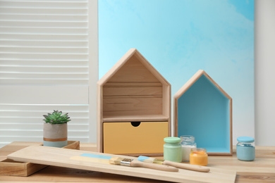 Photo of House shaped shelves, brushes and jars of paints on table indoors