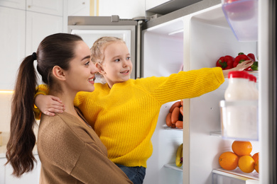 Young mother with daughter taking cucumber out if refrigerator in kitchen