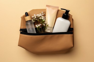 Preparation for spa. Compact toiletry bag with different cosmetic products and flowers on light brown background, top view
