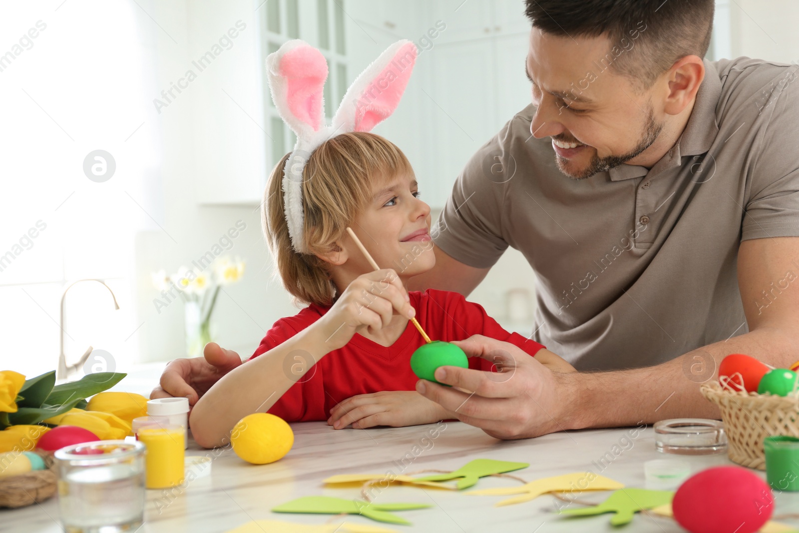 Photo of Happy son with bunny ears headband and his father painting Easter egg at table in kitchen