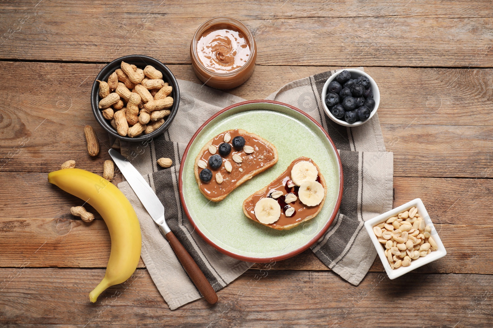 Photo of Toasts with tasty nut butter, banana slices, blueberries and peanuts on wooden table, flat lay