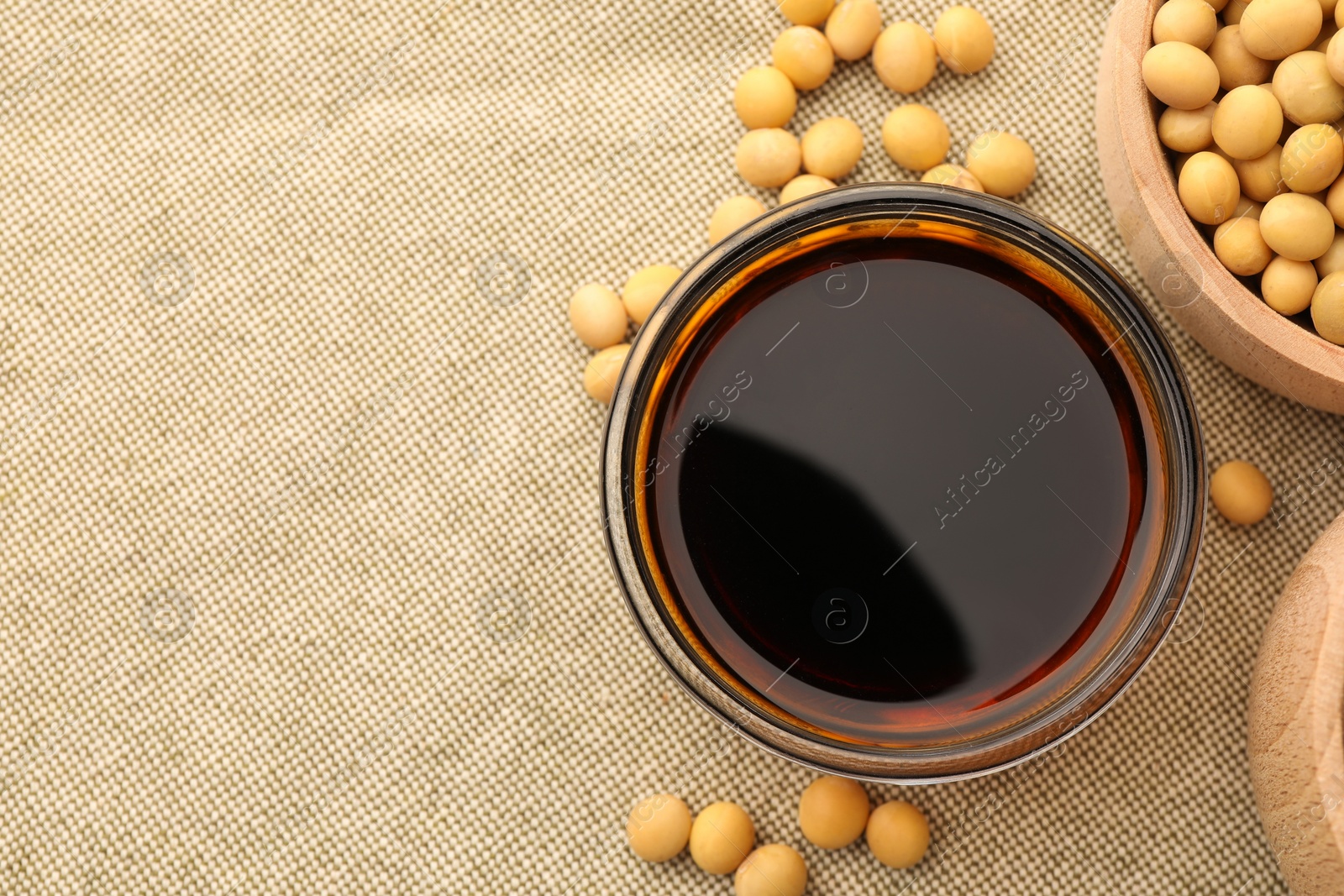 Photo of Soy sauce in bowl and soybeans on cloth, flat lay. Space for text