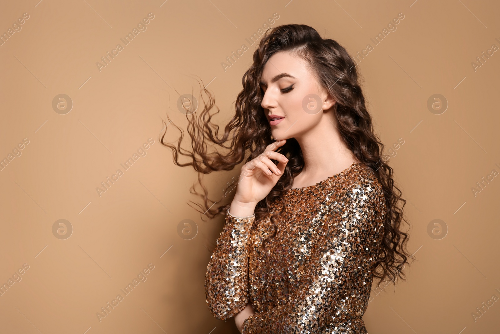 Photo of Beautiful young woman with long curly brown hair in golden sequin dress on beige background, space for text