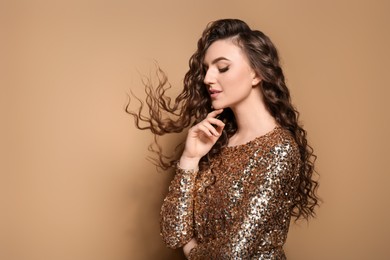 Beautiful young woman with long curly brown hair in golden sequin dress on beige background, space for text