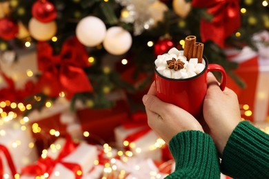 Woman holding cup of delicious cocoa with marshmallows, cinnamon sticks and anise star near Christmas tree, closeup. Space for text