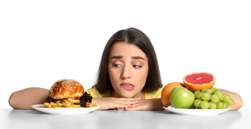 Photo of Doubtful woman choosing between fruits and burger with French fries on white background