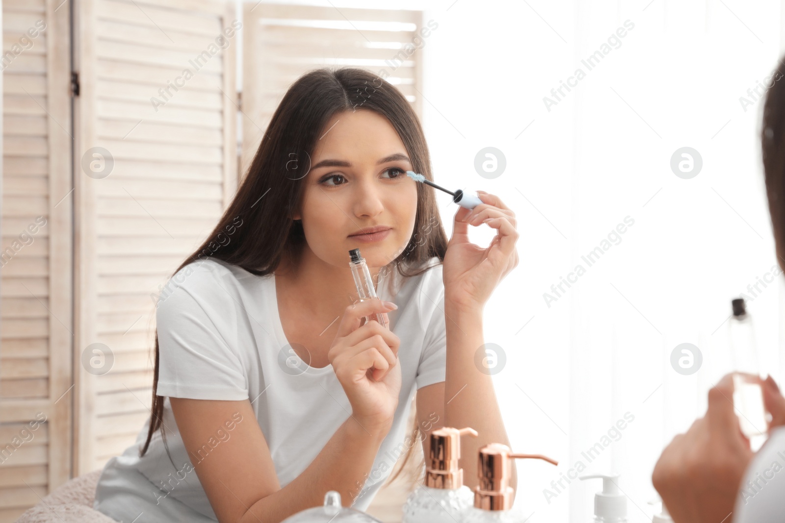 Photo of Beautiful woman applying oil onto her eyelashes near mirror indoors