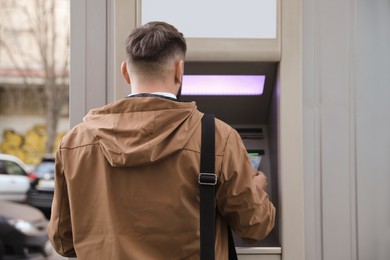Photo of Young man using cash machine for money withdrawal outdoors, back view