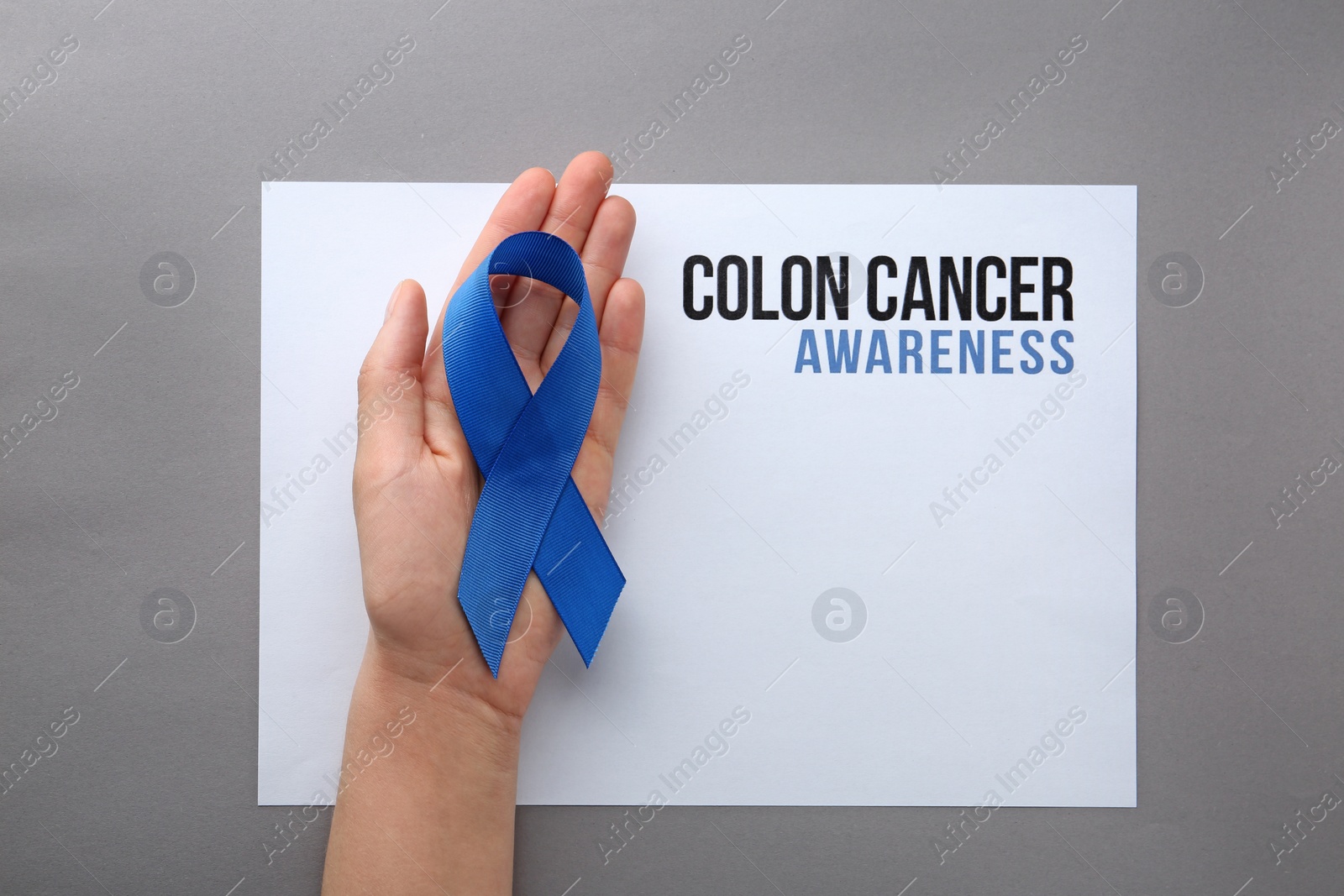 Photo of Woman holding blue ribbon near card with words COLON CANCER AWARENESS on grey background, top view. Space for text