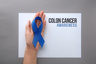 Photo of Woman holding blue ribbon near card with words COLON CANCER AWARENESS on grey background, top view. Space for text