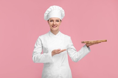 Happy chef in uniform holding empty wooden board on pink background