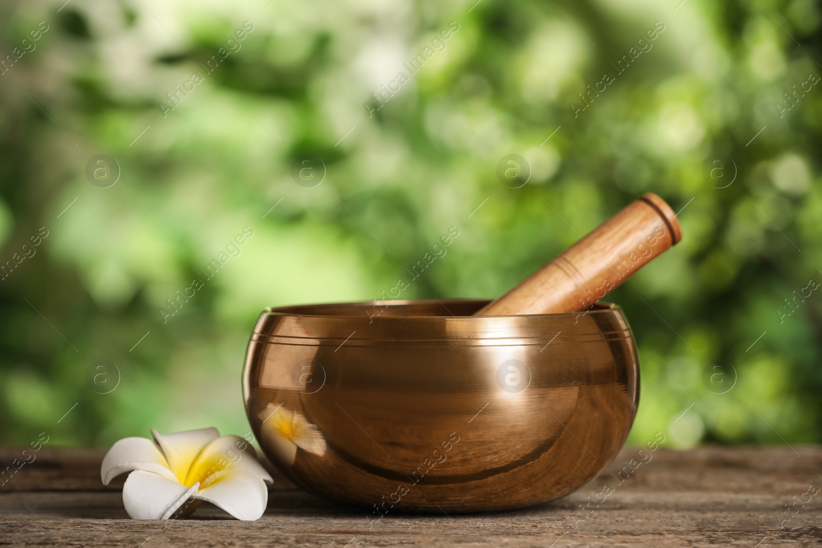 Photo of Golden singing bowl, mallet and flower on wooden table outdoors