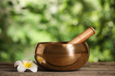 Photo of Golden singing bowl, mallet and flower on wooden table outdoors