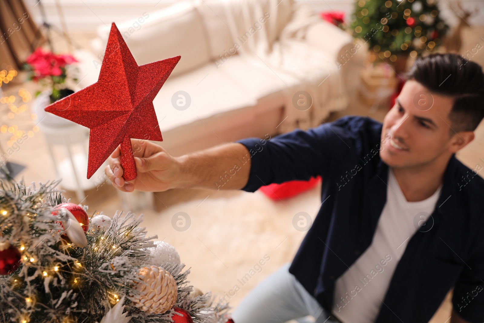 Photo of Man decorating Christmas tree in room, focus on star topper