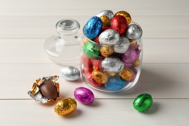 Glass jar with chocolate eggs wrapped in colorful foil on white wooden table