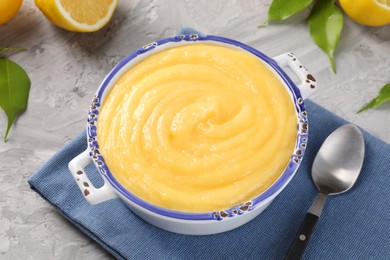 Photo of Delicious lemon curd in bowl, fresh citrus fruit, green leaves and spoon on grey table